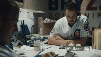 a man wearing a cubs jersey sits at a desk with a mlb.com logo in the corner
