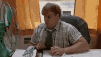 a man in a plaid shirt and tie is sitting at a desk in front of a green fan .