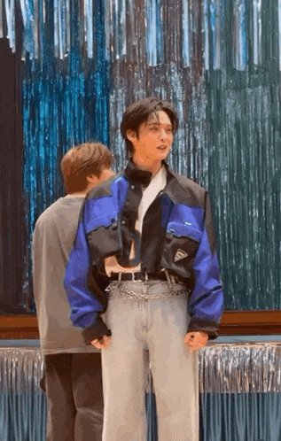 a man in a blue jacket and gray pants is standing next to another man in front of a blue curtain .