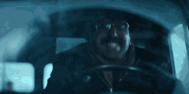 a man with a mustache and glasses is driving a car in the dark .