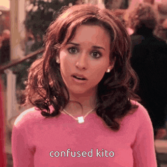 a woman is wearing a pink sweater and a necklace and is looking at the camera with a surprised look on her face .