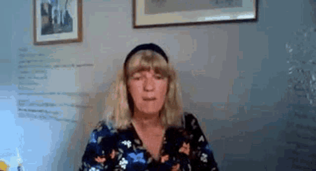 a woman in a blue floral shirt is sitting in front of a wall with a picture on it .