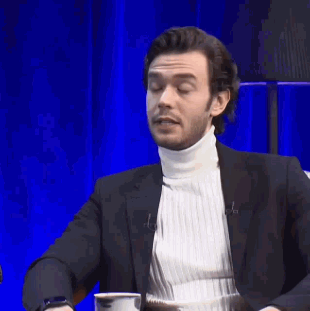 a man wearing a white turtleneck and a black jacket talks