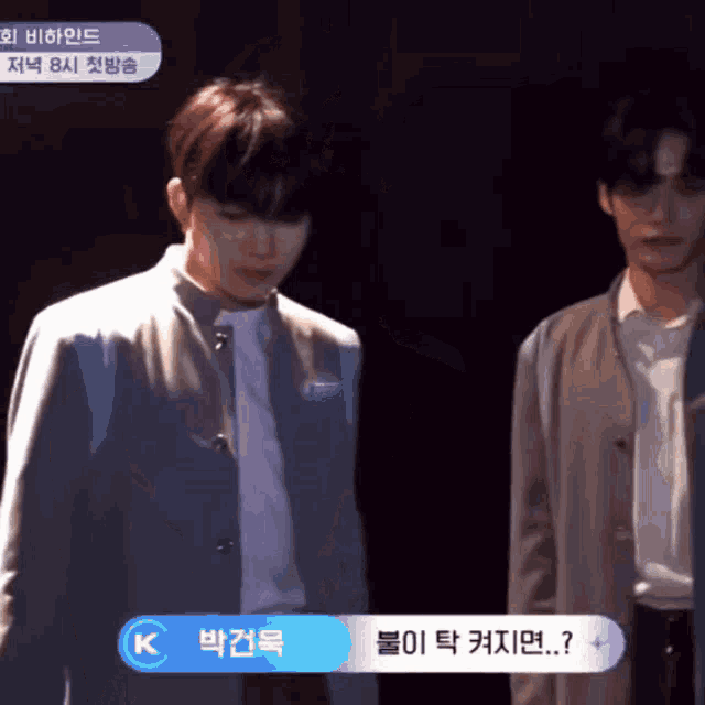 a man in a suit stands next to another man in a white shirt with a blue button that says k