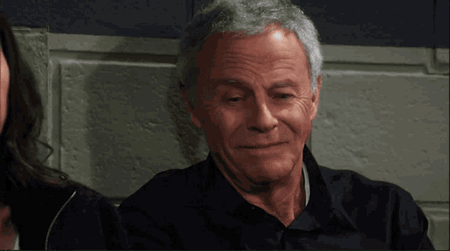a man with gray hair and a black shirt is sitting in front of a brick wall