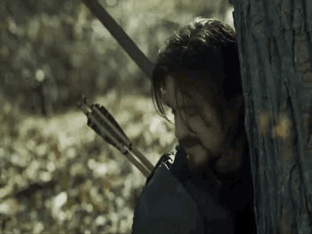 a man with a beard is holding a bow and arrow behind a tree .