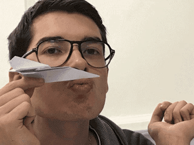 a young man wearing glasses is holding a paper airplane in front of his mouth