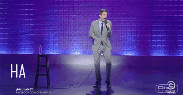 a man in a suit stands on a stage holding a microphone and says ha ha ha ha ha