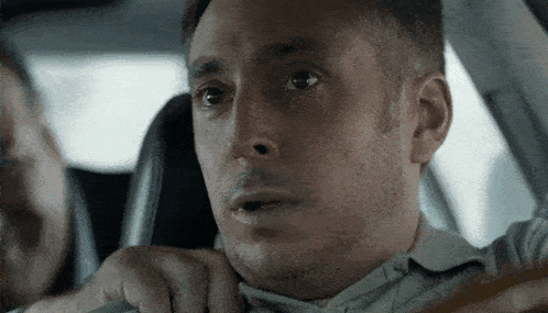 a man is sitting in the driver 's seat of a car and making a surprised face .