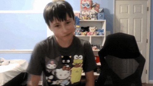 a young boy wearing a hello kitty t-shirt is standing in front of a chair