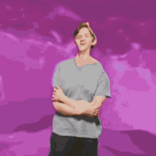 a man in a grey shirt is standing with his arms crossed in front of a purple background