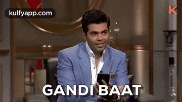 a man in a suit is sitting in a chair holding a cell phone and says gandi baat .