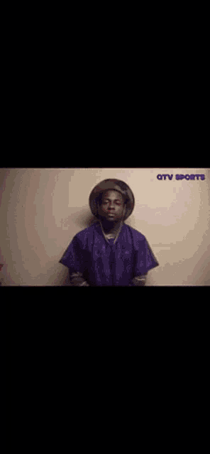 a man wearing a hat and a purple shirt is standing in front of a tv screen .