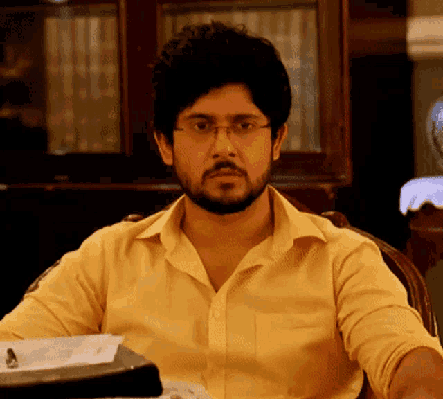 a man wearing glasses and a yellow shirt sits at a desk