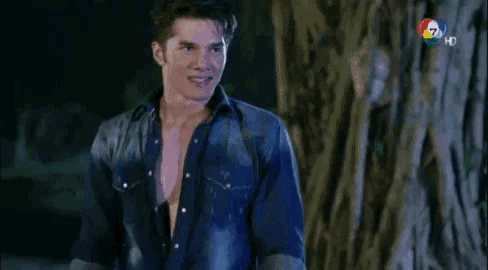 a man in a denim shirt is standing next to a tree at night .