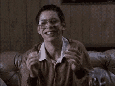 a man wearing glasses is sitting on a couch with his hands in the air and smiling .