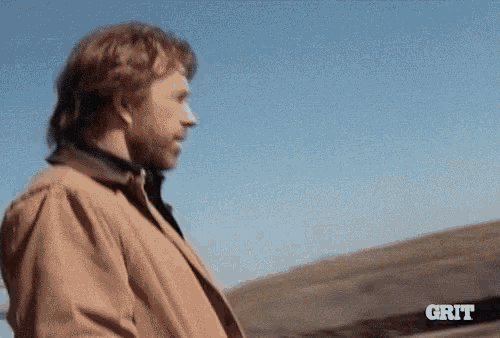a man in a brown jacket is standing on top of a hill with the word grit on the bottom .