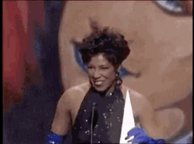 a woman in a black dress and blue gloves is standing in front of a microphone holding an award .