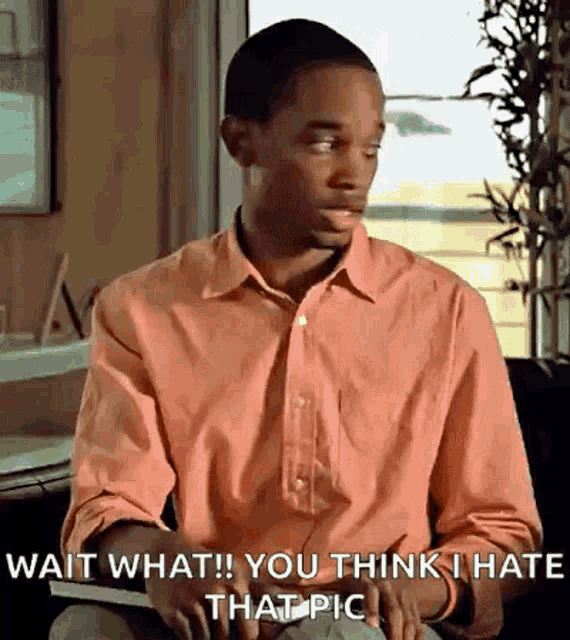 a man in a pink shirt is sitting in front of a window and says wait what you think i hate that pic .