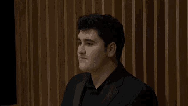 a man in a black suit stands in front of a wood paneled wall