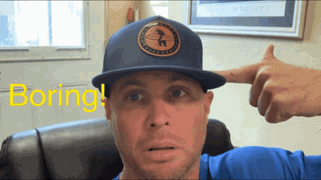 a man wearing a blue hat is pointing at his head with the word boring in yellow behind him