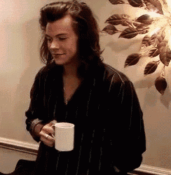 harry styles is holding a cup of coffee in front of a wall .
