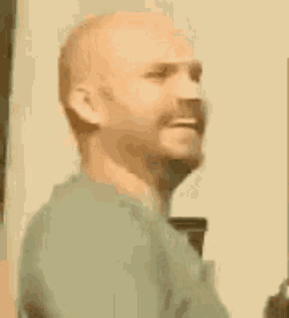 a bald man with a beard is standing in front of a wall and holding a gun .