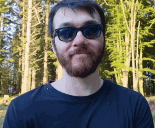 a man wearing sunglasses and a black shirt stands in front of trees