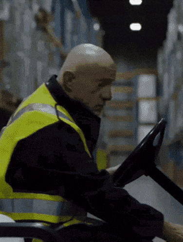 a bald man in a yellow vest is driving a forklift in a warehouse