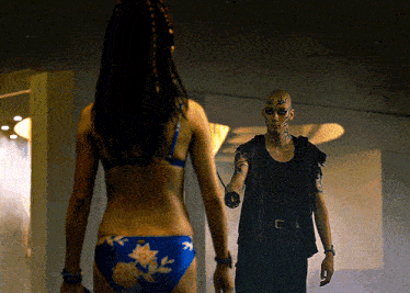 a woman in a bikini stands in front of a man with a face painted like a snake