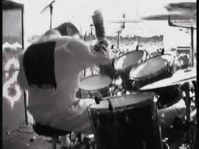 a man in a white shirt is playing drums in front of a wall with the letter s painted on it