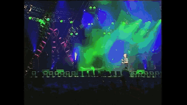 a man stands on a stage with a green light behind him and a crowd behind him