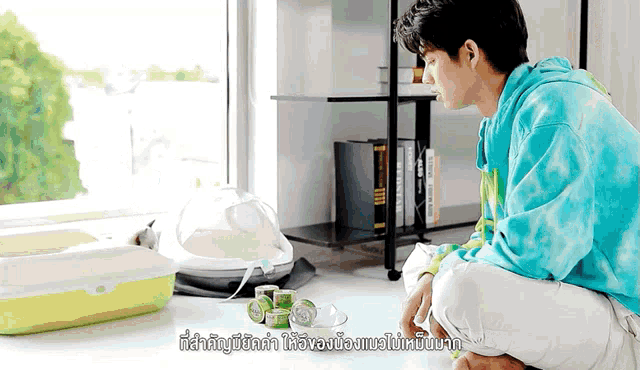 a man in a blue tie dye hoodie is kneeling on the floor in front of a litter box