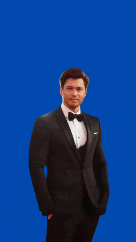 a man in a tuxedo with a bow tie stands in front of a blue background