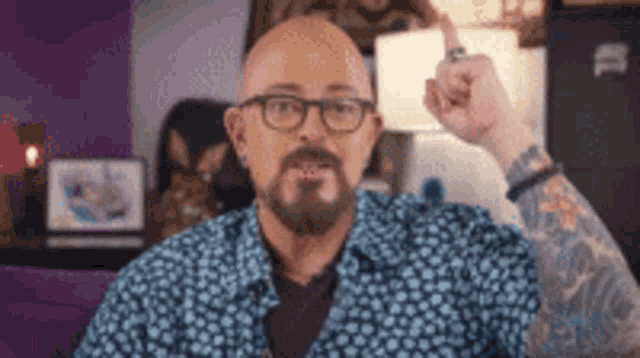 a bald man with glasses and a beard is sitting on a couch holding his finger up .