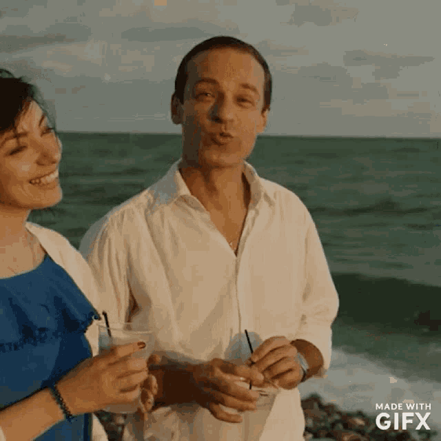 a man blowing a kiss while holding a drink next to a woman on the beach