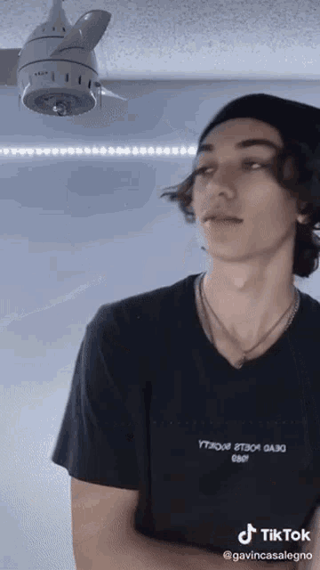 a young man wearing a black t-shirt that says tiktok stands in front of a ceiling fan