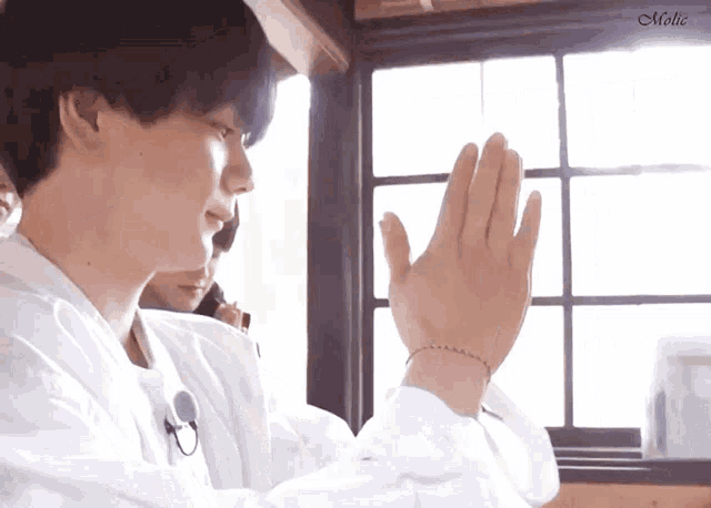 a man in a white shirt is praying in front of a window with his hands folded in front of him