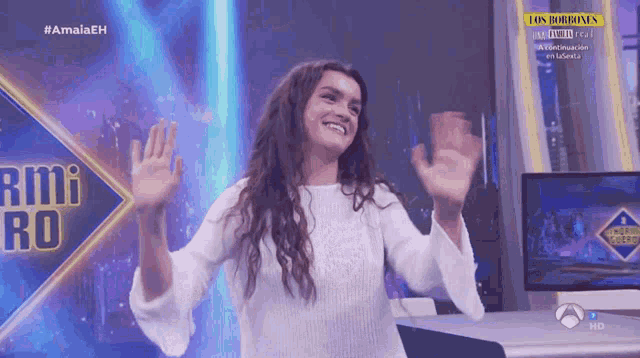 a woman in a white sweater is waving in front of a screen that says los bombones