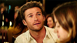 a man with a beard and a white shirt is talking to a woman in a crowded room .