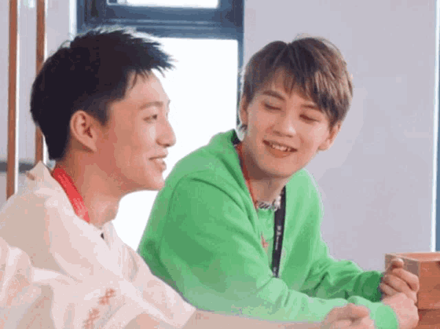 two young men are sitting next to each other and smiling . one is wearing a green sweater .