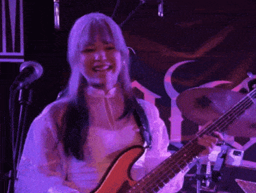a woman with purple hair is holding a bass guitar and singing into a microphone
