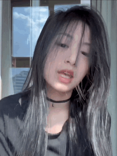 a woman with long gray hair and a choker is standing in front of a window and looking at the camera .