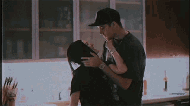 a man and a woman are kissing in a kitchen . the woman is wearing a hat .