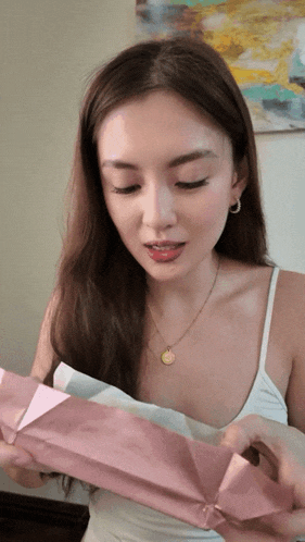a woman wearing a white tank top and a gold necklace is opening a pink box
