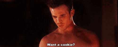 a shirtless man is holding a cookie in his hand and asking if he wants a cookie .