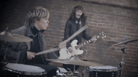 a man playing drums and a woman playing a guitar in front of a brick wall