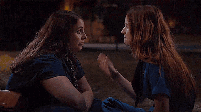 two women are sitting next to each other in the grass and touching their faces .