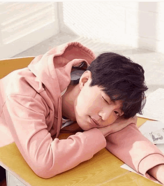 a young man in a pink hoodie is sleeping on a wooden desk