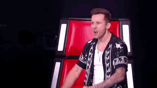 a man in a black and white shirt is sitting in a red chair and making a funny face .